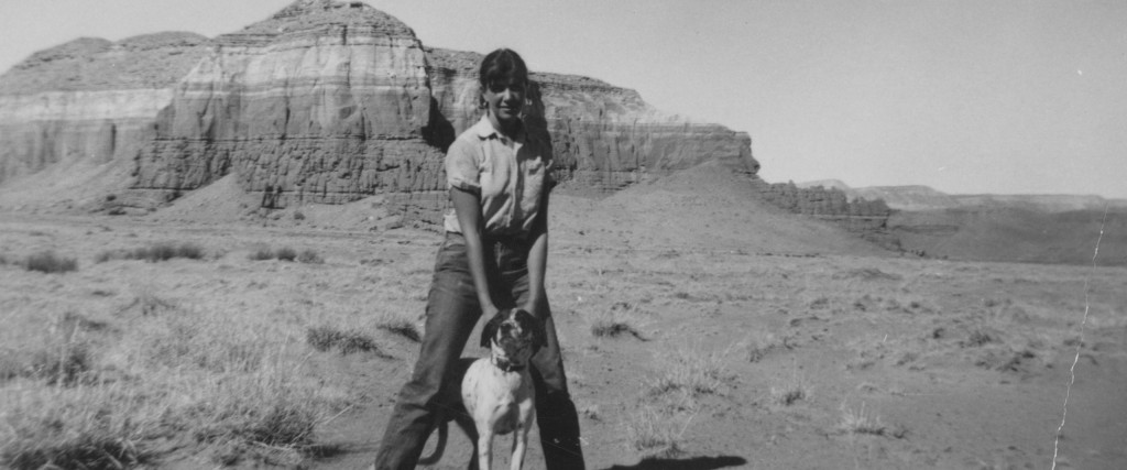 Foreground is a smiling woman (Rita Deanin Abbey) standing in a Utah desert mountain landscape background. In between her legs is a medium-sized dog (Homer). Both are looking at the camera.