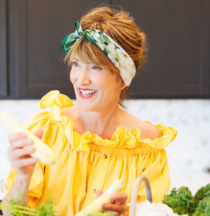 A woman (JoJami Tyler) is seen in her kitchen from the waist up, smiling with a basket of assorted vegetables and white flowers. She is holding a summer squash in each hand and looking off camera. Her orange hair is tied up and she is sporting a green bandana with white flowers in her hair.
