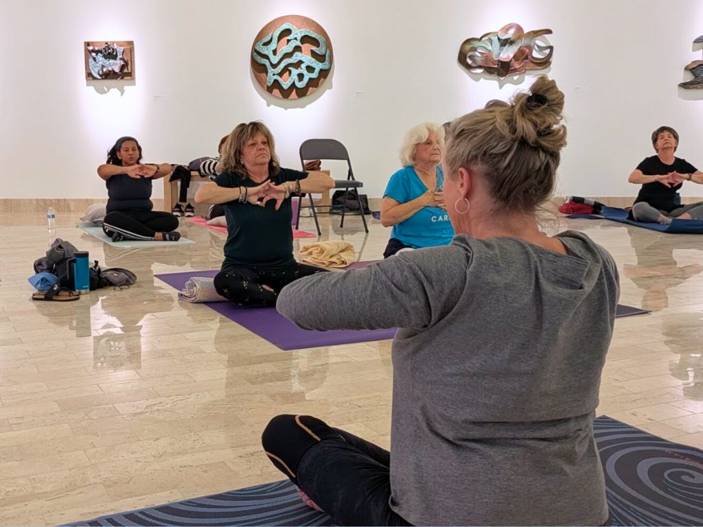 In foreground is Maureen Heher Karaffa leading a class of students, sitting on a purple yoga mat with her legs crossed and elbows out with her fingers clasped at her chest. The class in the background is mimicking her pose. The class is in the Wall of Creation gallery, so the students are surrounded by art.