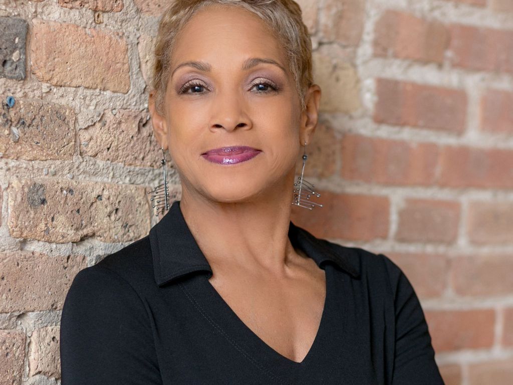 Headshot of a confident, smiling woman (Beth Burns) in a black shirt against a brick wall. She is the instructor for Women's Financial Literacy Empowerment.