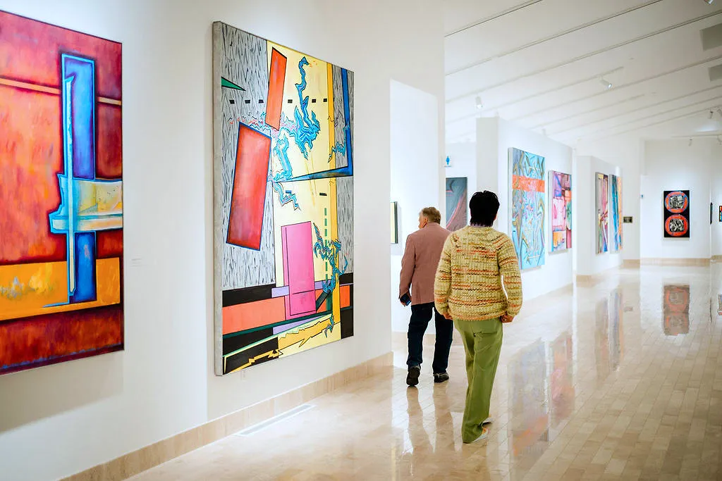 Two guests in the center of the photo walk the hallway of the main gallery of the museum, which features five visible, colorful, large-scale paintings. Their backs are to the camera, but you can see their heads are turned to view the paintings which are all abstracts with rectangular and organic shapes ranging from blues, reds, and yellows as the main colors. The guest closest to the camera is wearing soft green pants with a matching yellow and green sweater.