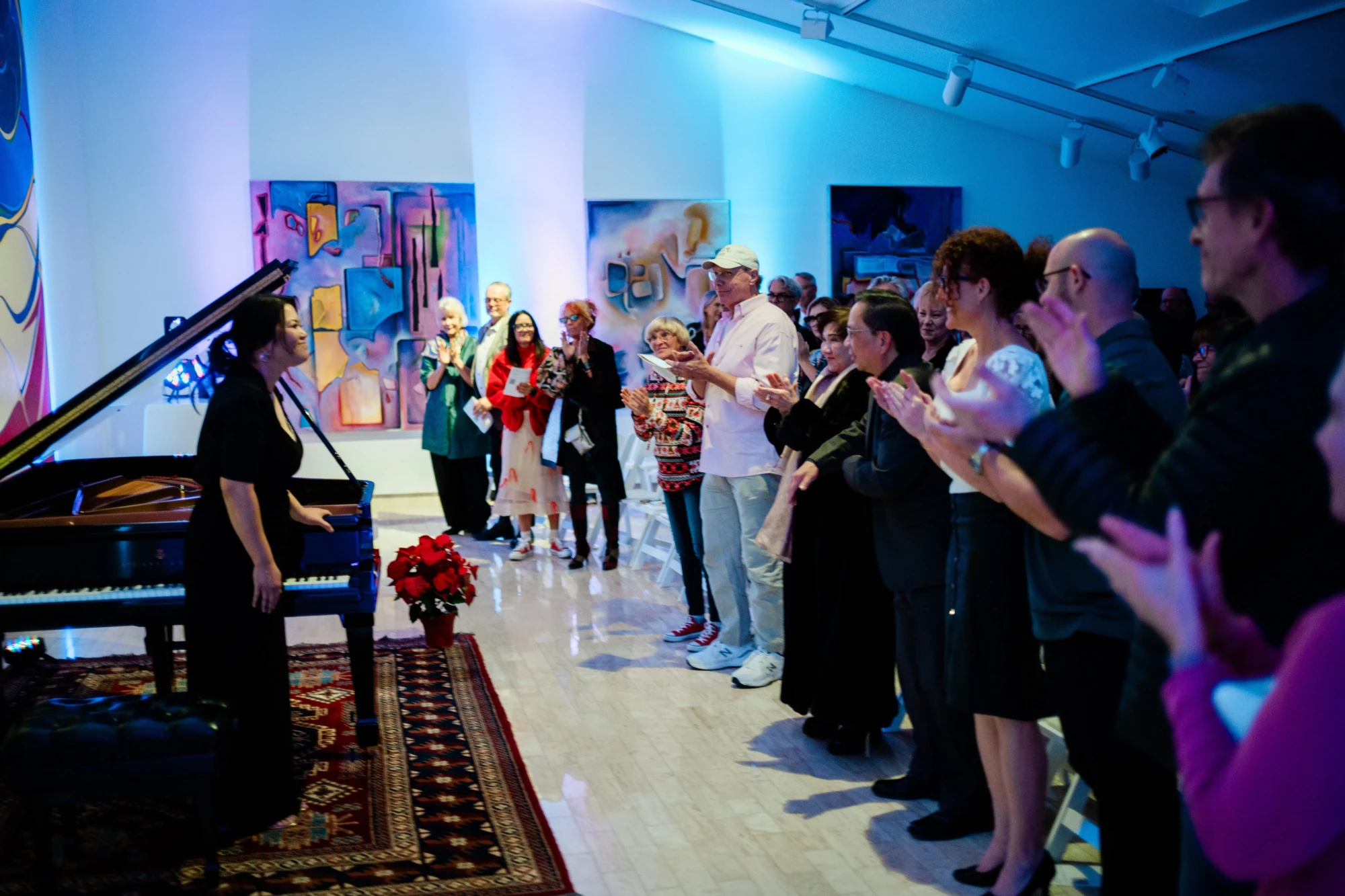 On the left the pianist stands, dressed in concert black and smiling in a small bow with her piano. On the right, the audience gives her a standing ovation. The room is lit by blue lighting, giving it a blue tone.