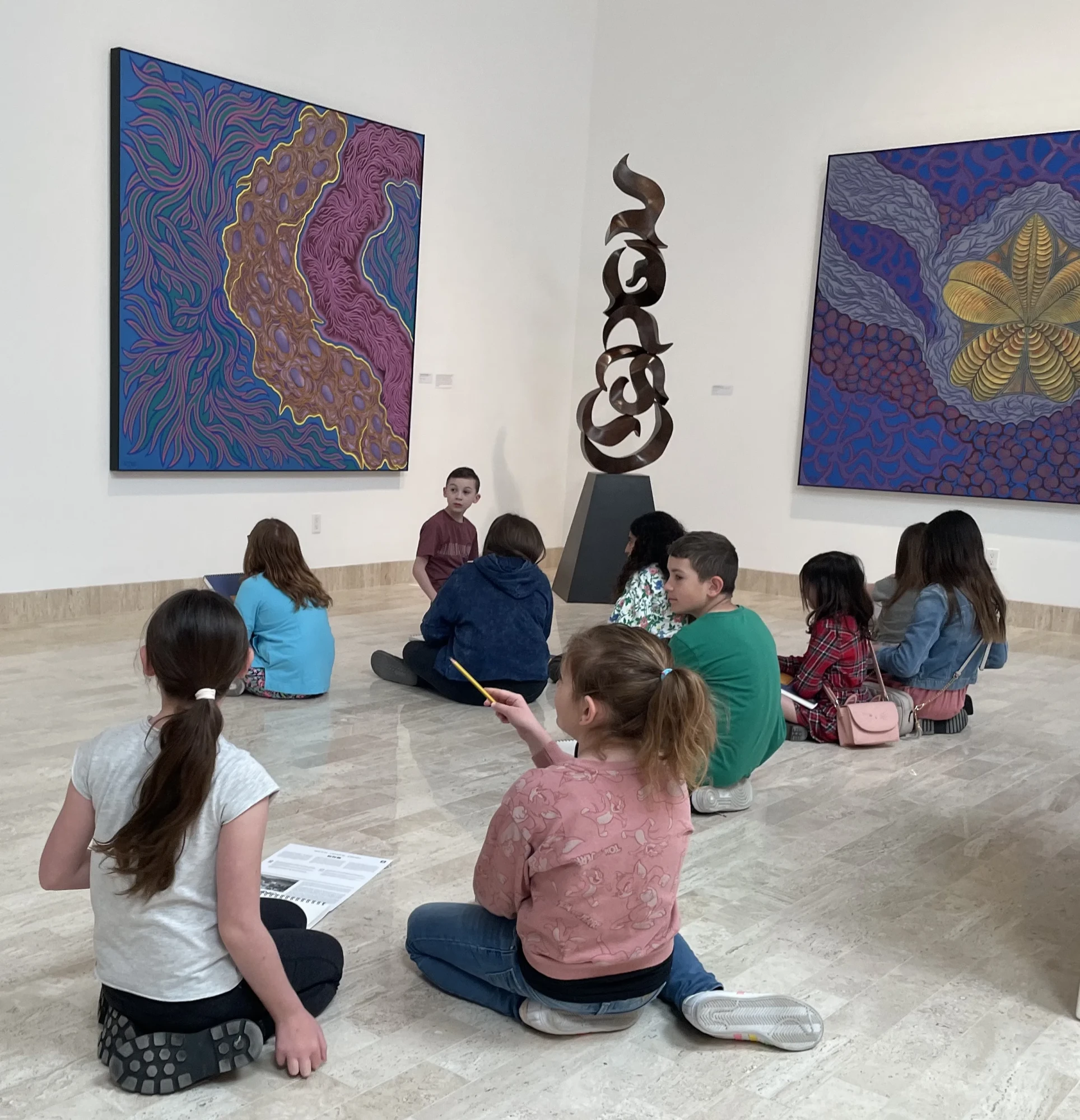 Ten children sit on the floor of the main gallery, each with sketchbooks, sketching the artwork. Two girls in the foreground have their heads turned toward an off-camera piece. The one on the right has her pencil pointing at the piece. The children in the background are all looking foreword, save two which are engaged in conversation. There are three pieces which can be seen (from left to right): a purple, yellow, and pink sea life looking piece, a tall sculpture of bronze, ancient-looking symbols stacked on top of each other, and a painting with a yellow center, which unfolds to reveal a pomegranate-esque scene.