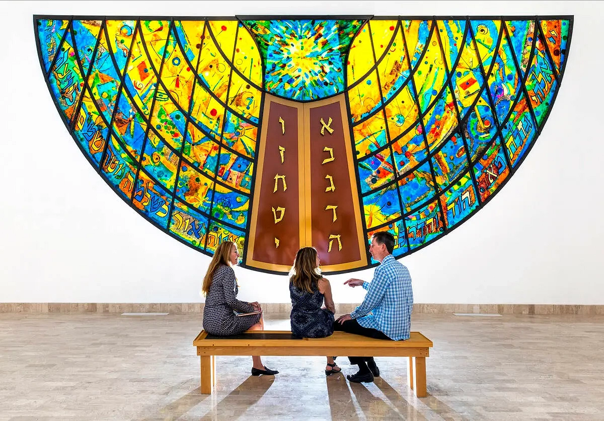 A photo of Wall of Creation, a stained glass mural created by Rita Deanin Abbey. It is a half circle shape, rooted in the center by two thin, rounded doors with Hebrew letters. Above the doors are two hands holding a flaming heart. From the center and out, the piece fades from yellow to blue, mimicking the sun in the sky. The piece also has Hebrew letters on the bottom edges that curve with the piece. Finally, different shapes and swirls on the glass make the piece feel full.