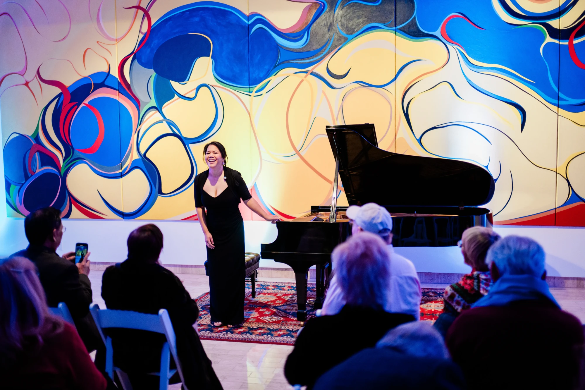 A smiling woman (Alexandria Le) stands, mid bow next to a piano to receive a clapping audience. The lighting is soft and blue, lighting up the yellow and blue mural, Bridge Mountain, by Rita Deanin Abbey.