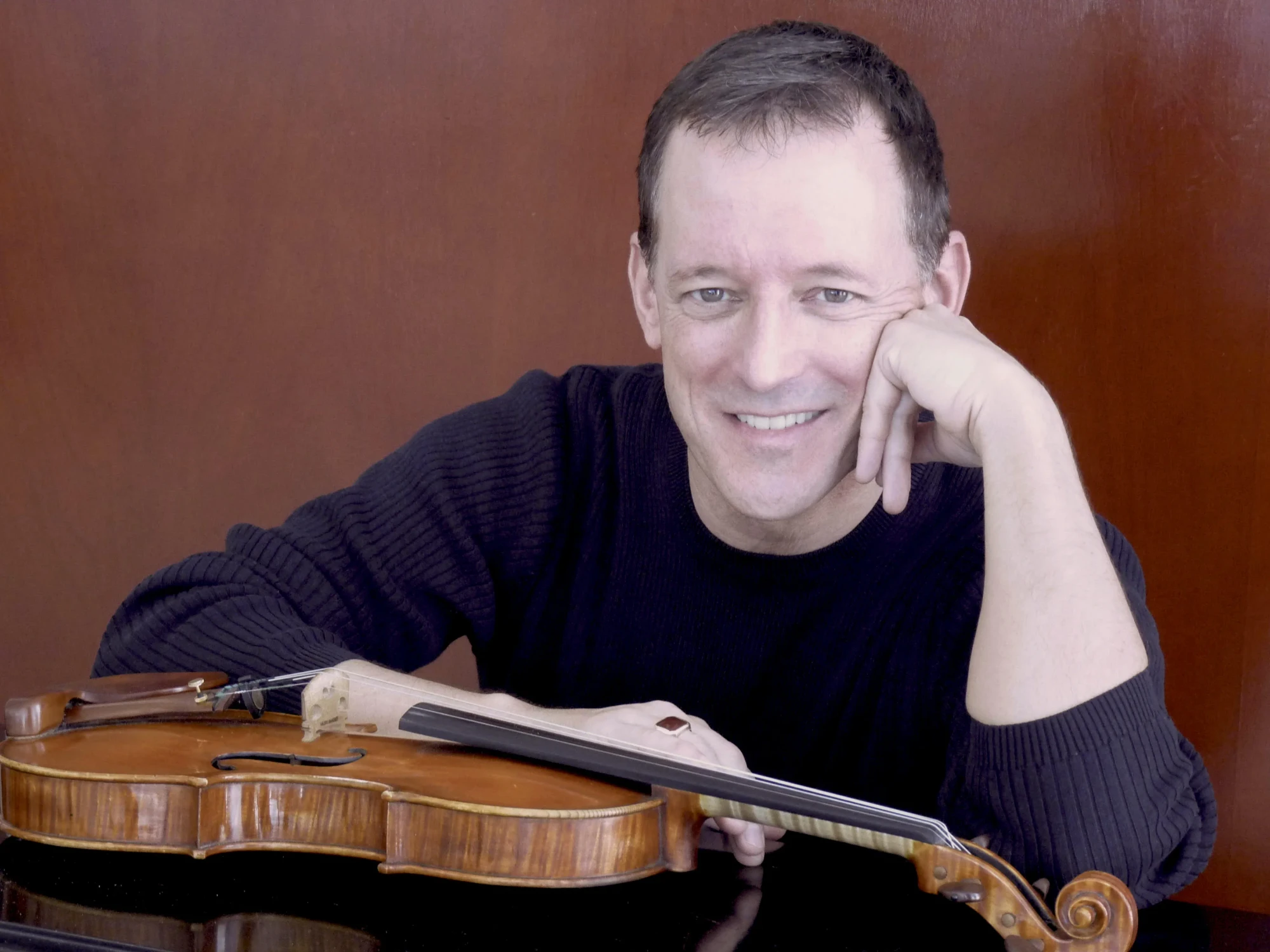A smiling man, Robert Taylor, seated for a portrait photograph. His violin is layed in front of him.