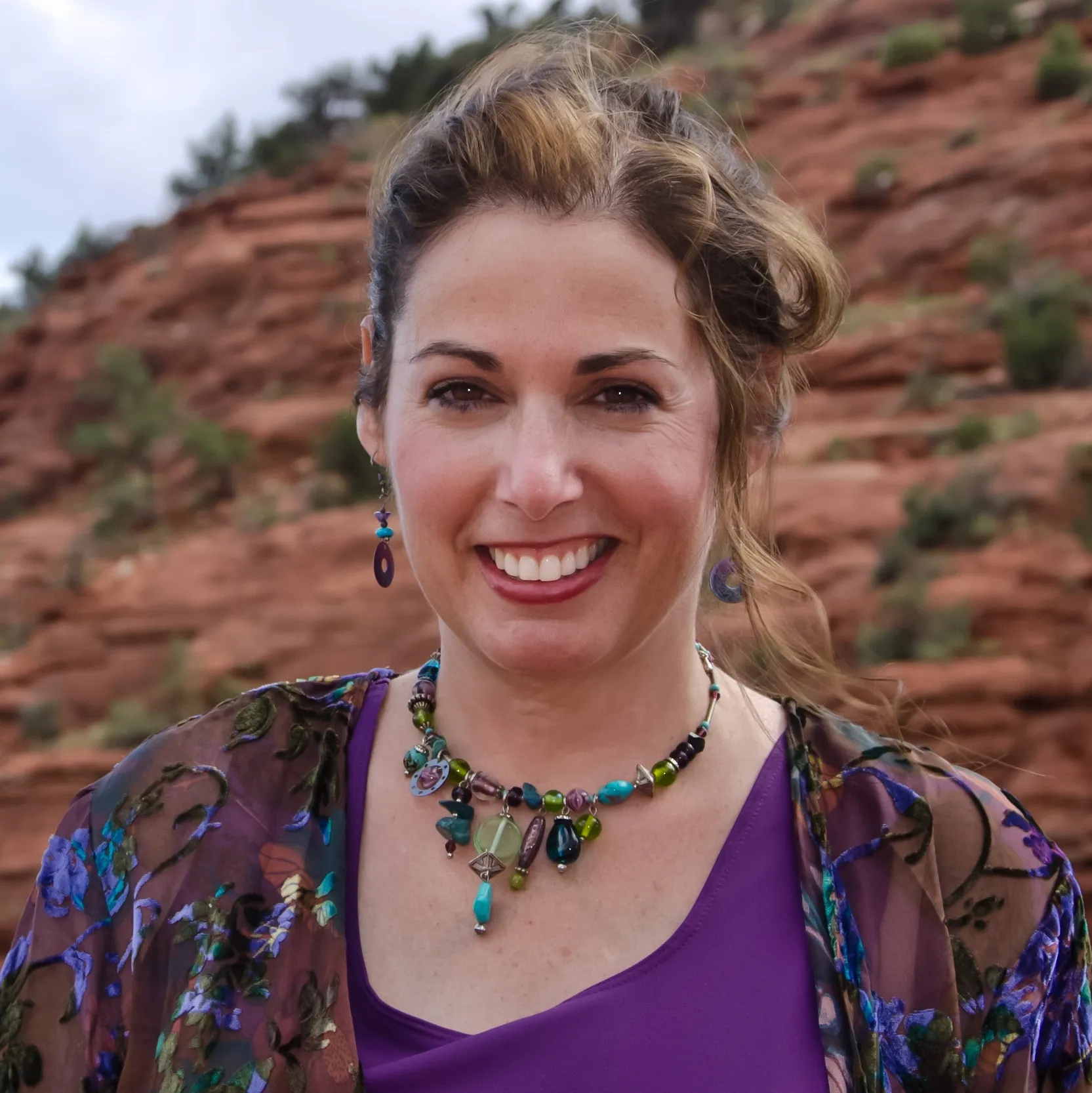 Headshot of a woman in the mountains, smiling at the camera and wearing a professional, purple top and flower jacket.