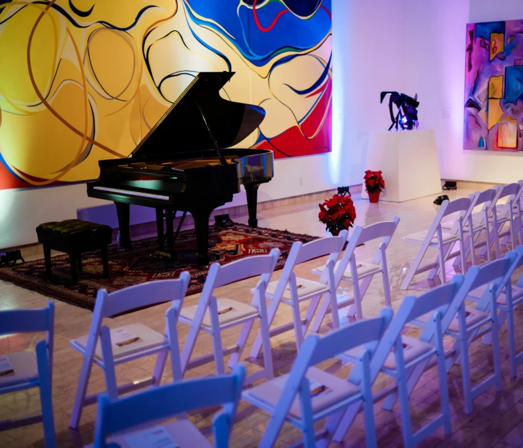 A piano set up for a concert in fron of 'Bridge Mountain' by Rita Deanin-Abbey. Several chairs are aligned in a row to view the show. A flower arrangement can be seen by the piano.