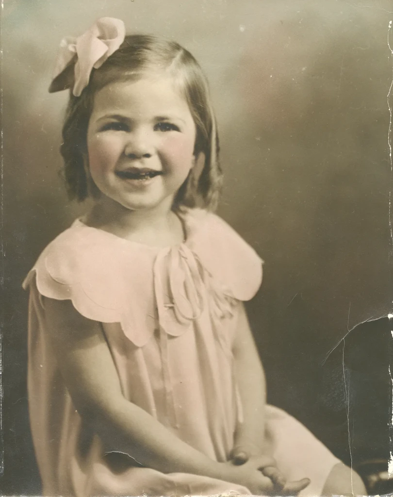 A young toddler smiles at the camera wearing a pink bow in her curled hair and a pink dress with a scalloped collar. She is posed with her hands in her lap. This old photo has been slightly colorized.