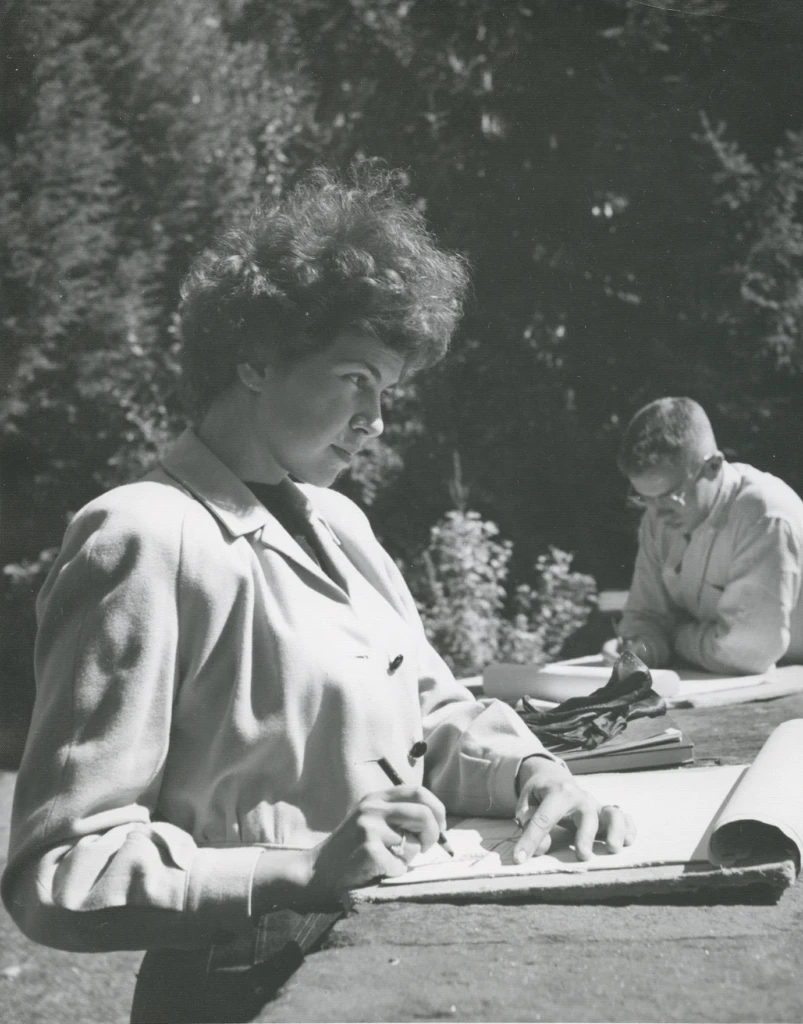 Rita stands outside in an art class working on her sketch book. She has a determined look of inspiration in her eyes while sketching something. In the background, another student sketches.