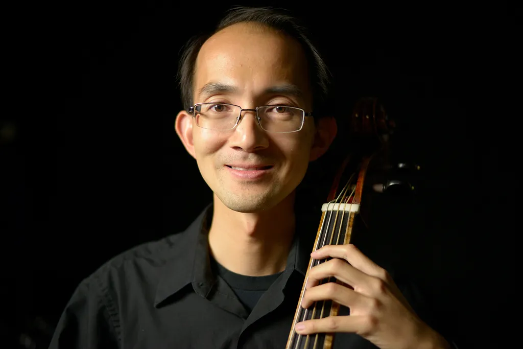 A smiling man, Ka-Wai Yu, with their baroque viola de gamba against a black background.