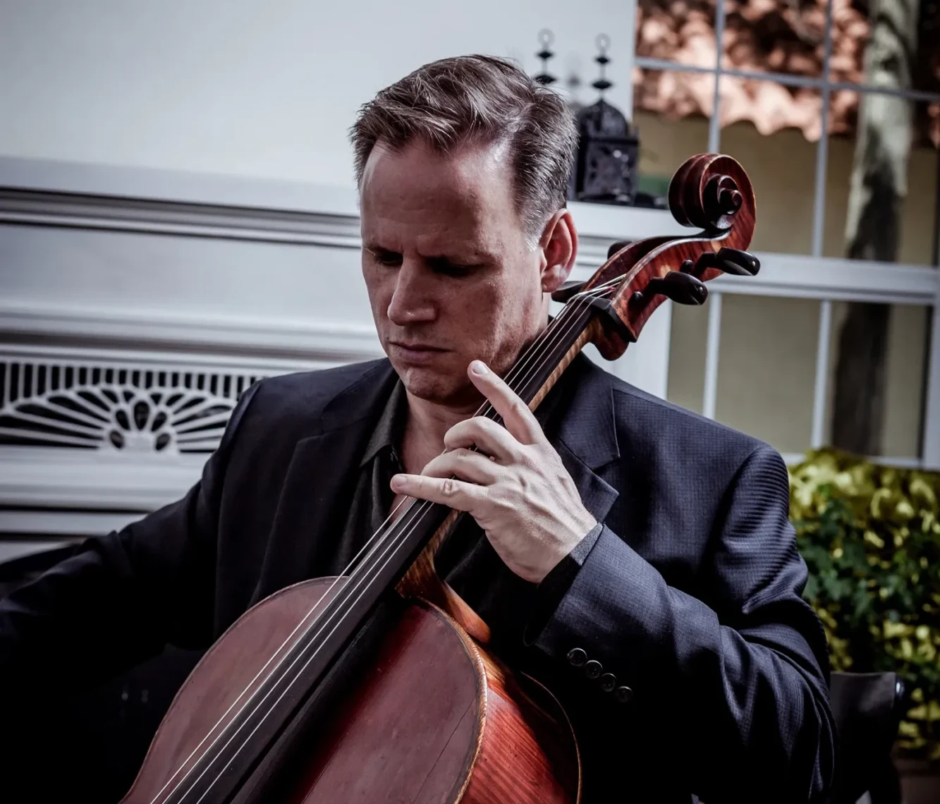 A serious man, Andrew Smith, playing the cello while in front of a fireplace.