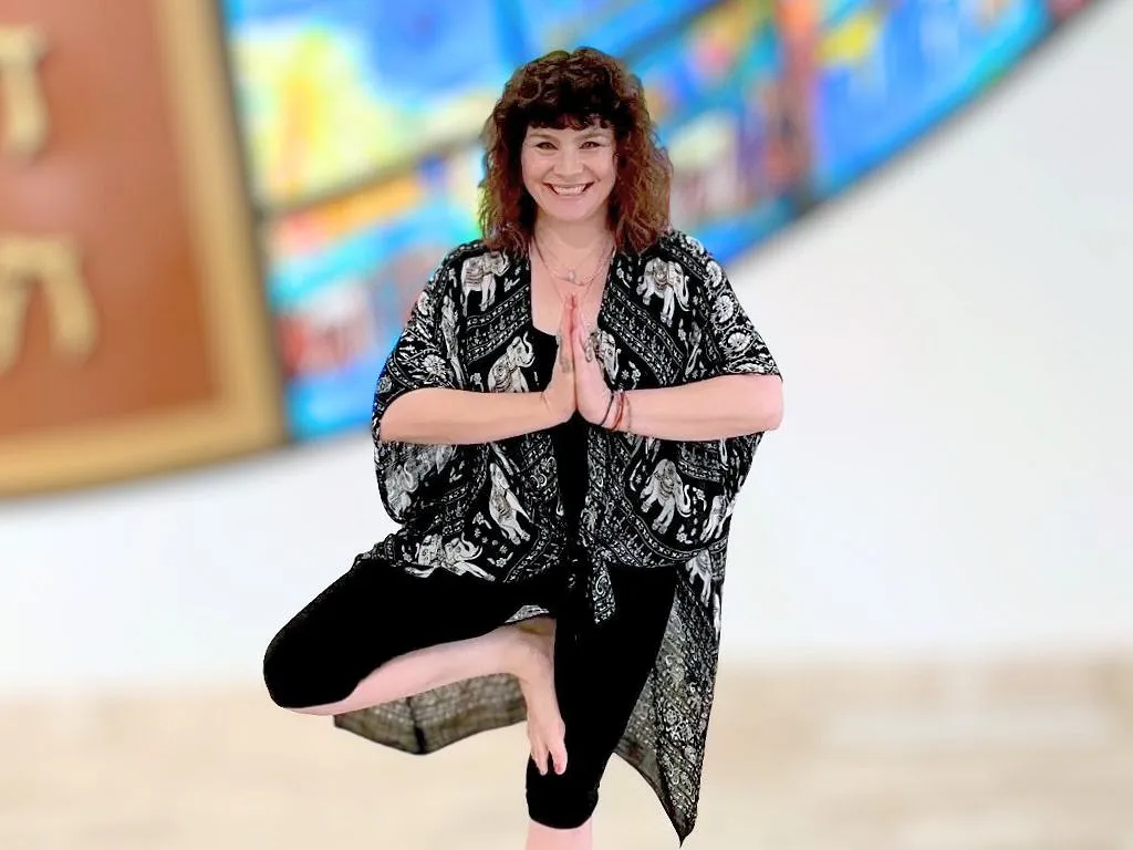A woman, Nicole Scheerer, smiling while performing the one-leg tree pose. She is in front of the Wall of Creation