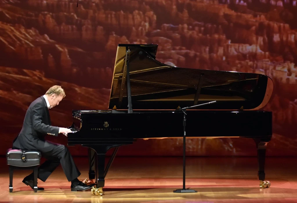 A man, Philip Fortenberry playing on a grand piano on a stage. A mountain range is projected as the background of the stage.