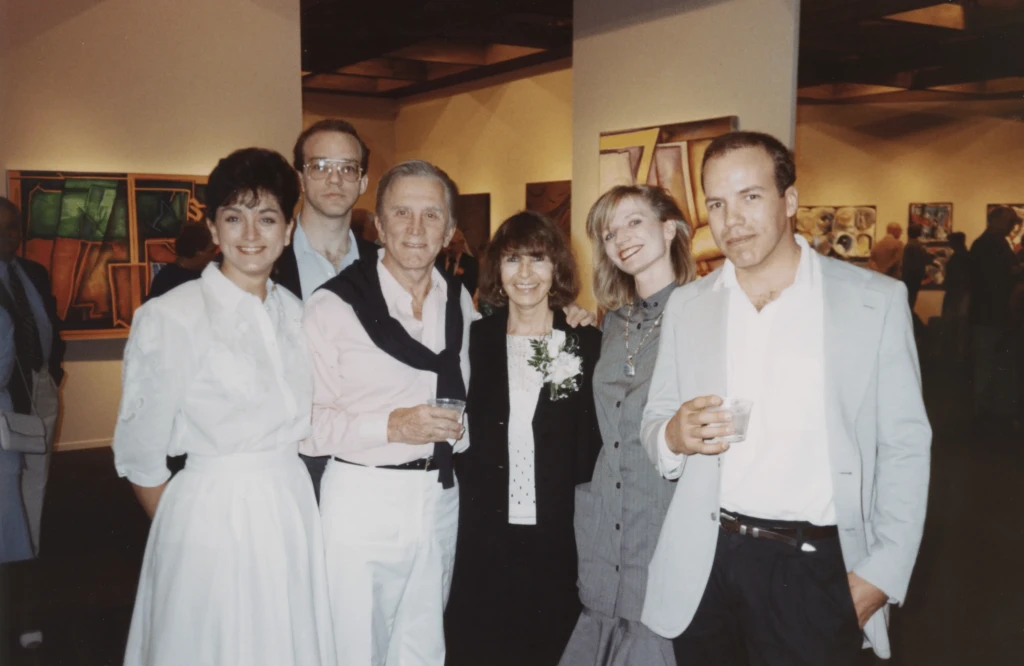 Six people stand in a gallery looking and smiling at the camera. In the center of the photo are Kirk Douglas and Rita Deanin Abbey. They are surrounded by Rita's sons, Aaron and Joshua, and the sons' wives.