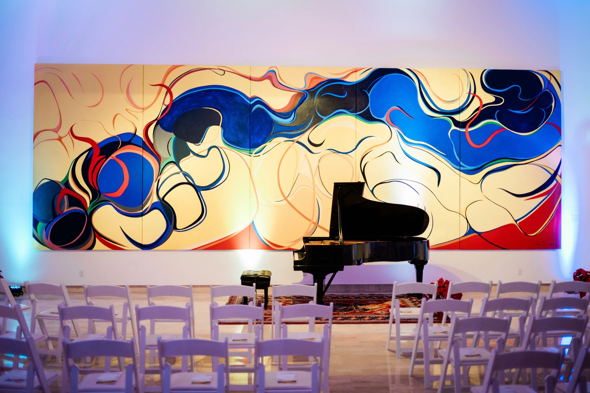 A piano set up for a concert in fron of 'Bridge Mountain' by Rita Deanin-Abbey. Several chairs are aligned in a row to view the show. A flower arrangement can be seen by the piano.