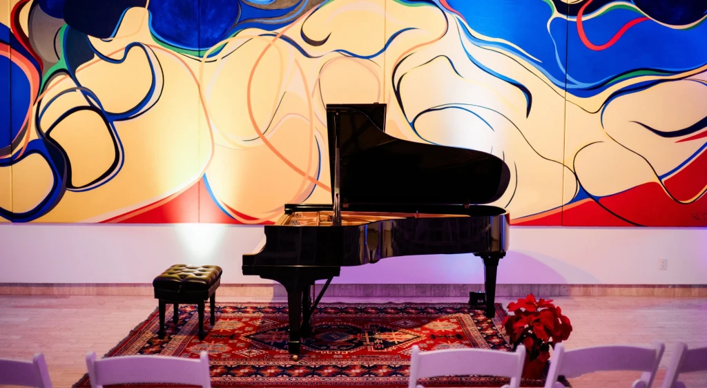 A piano set up for a concert in fron of 'Bridge Mountain' by Rita Deanin-Abbey. Several chairs are aligned in a row to view the show. A flower arrangement can be seen by the piano.