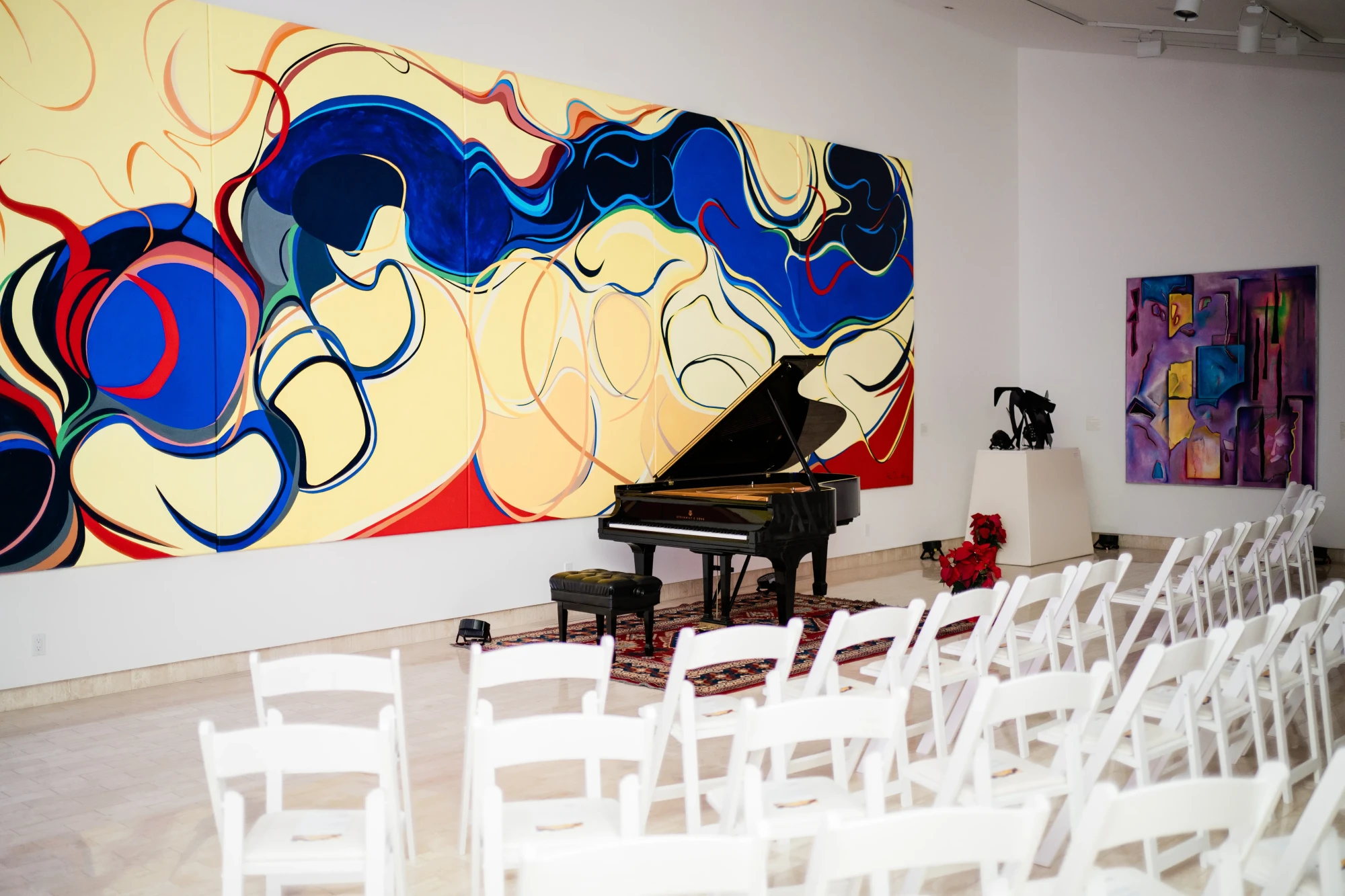 A piano set up for a concert in fron of 'Bridge Mountain' by Rita Deanin-Abbey. Several chairs are aligned in a row to view the show. A flower arrangement can be seen by the piano.