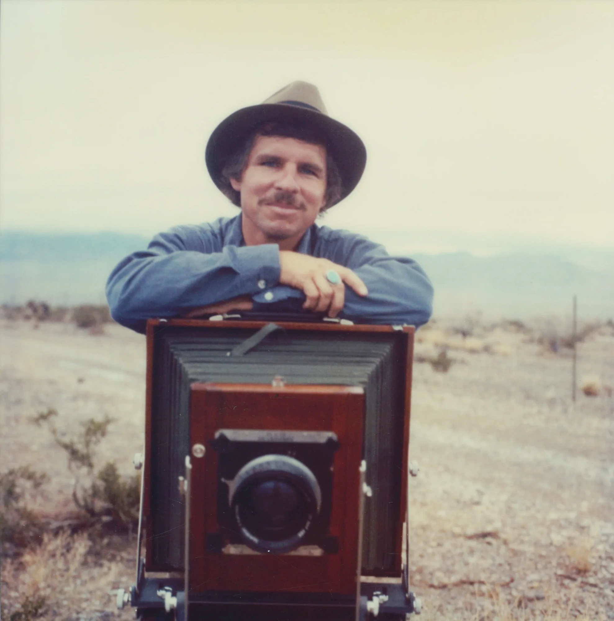 A man wearing a blue denim shirt and brown wide-brimmed hat resting his arms on a large photobox camera on a stand. Behind him is an expanse of yellowed desert landscape.