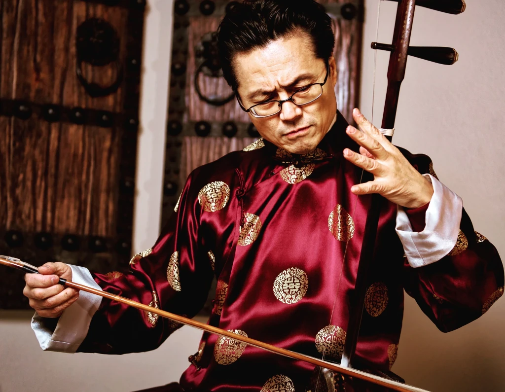Hong Wang wearing a shiny traditional shirt and playing an ancient instrument, look of deep concentration on his face